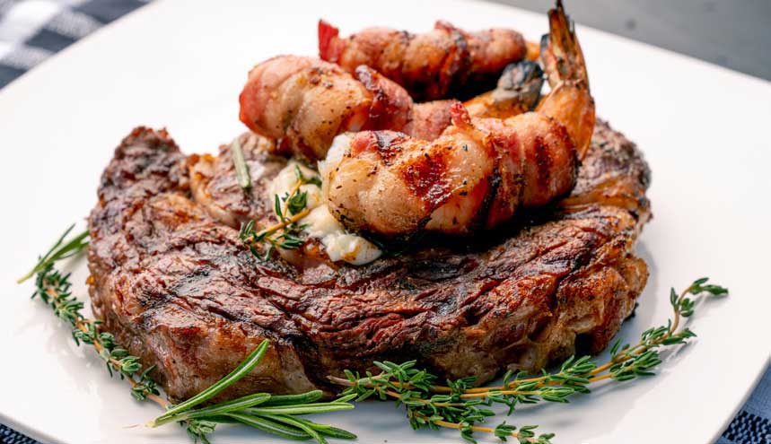 grilled shrimp resting on top of a steak, with rosemary decorating the plate
